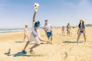 Group of multiethnic friends playing soccer on the beach - Football match on the sand on summertime - Tourists having fun on vacation with beach games